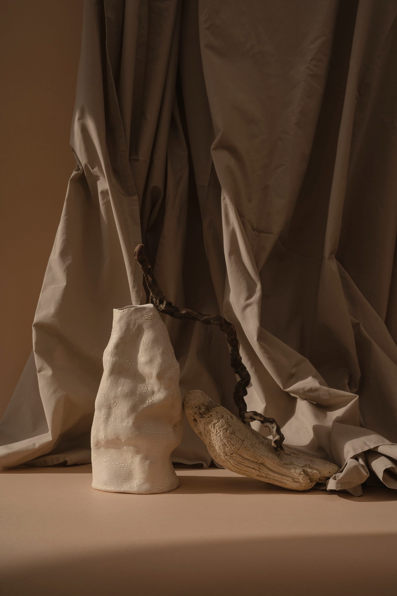 A ceramic vase beside a tree branch and draped fabric, in warm neutral tones